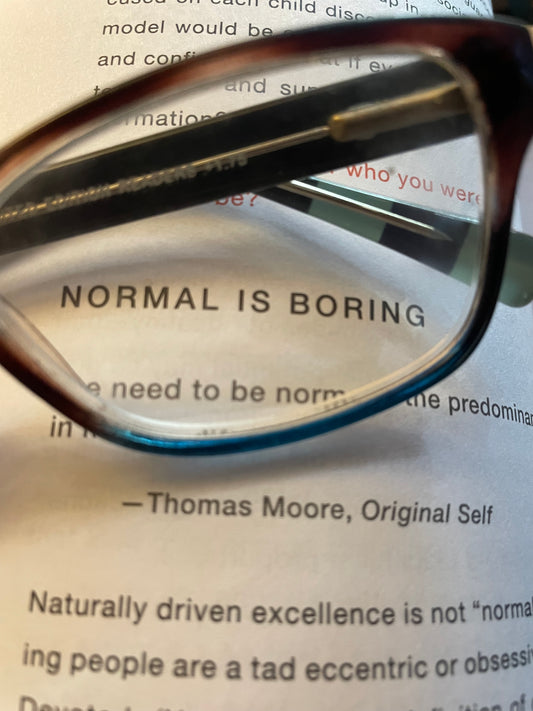 The lens of a pair of reading glasses surrounds the phrase Normal is Boring in a book.